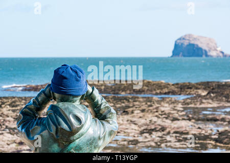 North Berwick, East Lothian, Schottland, UK, 23. März 2019. UK Wetter: sonnig Wetter im Frühling in der Stadt am Meer. Bronze Statue namens Watcher von Kenny Hunter mit einem warmen hat als er Umfragen Bass Rock gannet Kolonie durch ein Fernglas Stockfoto