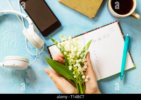 Frühling Blumen Lilien in weibliche Hände und Schreiben im Notebook, Smartphone auf das Tal und die Kopfhörer auf den Tisch aus Stein. Ansicht von oben flach Hintergrund wi Stockfoto