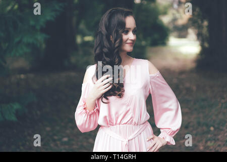 Süße weibliche junge Brünette mit Locken in ein rosa Kleid im Park, die Personifizierung der Zärtlichkeit und der Weiblichkeit Stockfoto