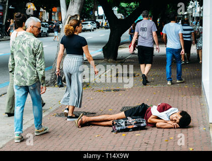 Rio de Janeiro, Brasilien, 23. März 2019: obdachlose junge Mann schlafen während Fußgänger neben ihm auf der belebten Fußgängerzone Bürgersteig in der Fülle zu Fuß