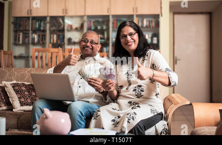 Ältere indische/asiatische Paar Buchhaltung, zu Hause finanzieren und Rechnungen mit Laptop, Rechner und Geld zu überprüfen, während auf Sofa/Couch zu Hause sitzen Stockfoto