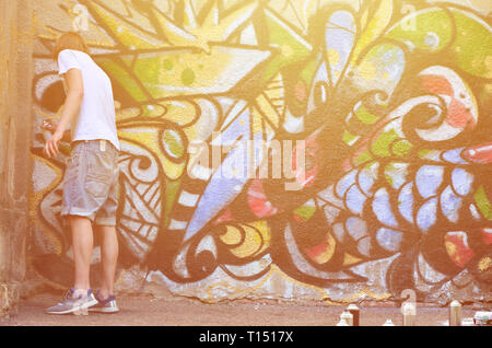 Foto von einem jungen Kerl in Denim Shorts und ein weißes Hemd. Der Mann stützt sich auf die Graffiti an der Wand eine Zeichnung mit Aerosol Lacke in verschiedenen Farben. Die anhand von quantitativen Simulatio Stockfoto