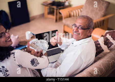 Ältere indische/asiatische Paar Buchhaltung, zu Hause finanzieren und Rechnungen mit Laptop, Rechner und Geld zu überprüfen, während auf Sofa/Couch zu Hause sitzen Stockfoto