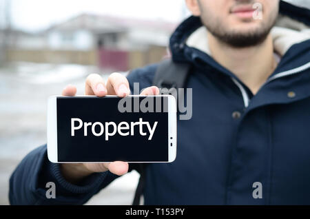 Ein junger Kerl zeigt eine Inschrift auf dem Smartphone Display auf dem Hintergrund von einem großen Haus im Winter. Eigenschaft Stockfoto