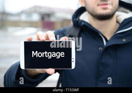 Ein junger Kerl zeigt eine Inschrift auf dem Smartphone Display auf dem Hintergrund von einem großen Haus im Winter. Hypothek Stockfoto