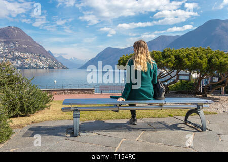 Eine schöne elegante Frau auf einer Parkbank und an den Luganer See und die Alpen in der Schweiz suchen Stockfoto