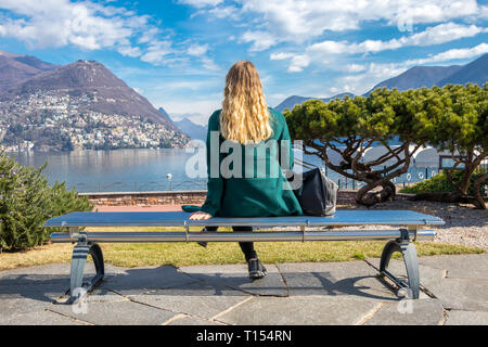 Schöne elegante Frau auf einer Parkbank und an den Luganer See und die Alpen im Tessin Kanton der Schweiz suchen Stockfoto