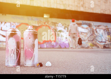 Mehrere verwendet, Spraydosen mit rosa und weisser Farbe auf den Asphalt gegen die ständigen Kerl vor einer bemalten Wand in farbigen Graffiti Zeichnungen Stockfoto