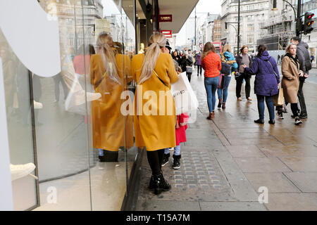 Rückansicht der Frau in modischer Gelbgold Wolle Mantel Reflexion in einem Schaufenster & Geschäfte auf Oxford Straße im Februar London Großbritannien KATHY DEWITT Stockfoto