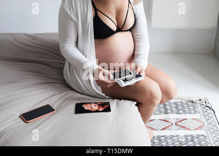 Schwangere Frau zu Hause sitzen auf dem Bett mit Ultraschall Bilder Stockfoto