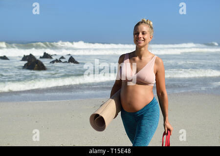 Aktive schwangere Frau gehen mit gymnastikmatte am Strand Stockfoto