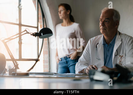 Ernsthafte leitender Arzt am Schreibtisch in der medizinischen Praxis sitzen mit Frau im Hintergrund Stockfoto