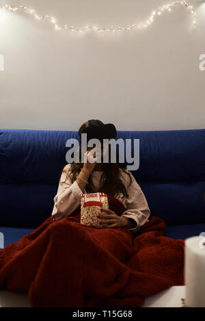 Junge Frau mit VR-Headsets essen Popcorn auf dem Sofa zu Hause in der Nacht Stockfoto