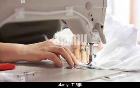 Weibliche Hände nähen weißen Stoff auf der Nähmaschine am Arbeitsplatz mit professionellen Schere für den Hintergrund. Näherin Hände halten Textil für Dres Stockfoto