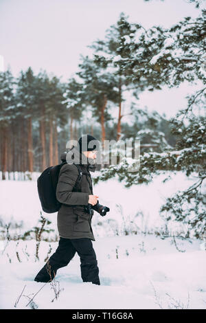 Junger Mann Backpacker mit Foto Kamera nimmt Fotos im Winter schneebedeckten Wald. Aktive Hobby. Wanderer Wandern in Verschneiten Pinienwald Stockfoto