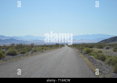 Eine unbefestigte Straße führt durch die felsige Mojave-Wüste in Kalifornien in ein trockenes Tal und weit entfernte Bergketten unter klarem blauen Himmel. Stockfoto