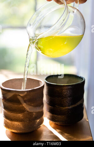 Nahaufnahme der Person gießen grüner Tee in Tassen im Sommer im Haus Frühstück mit gelben Sencha Farbe und Stream durch Fenster zwei japanische Teetassen Stockfoto