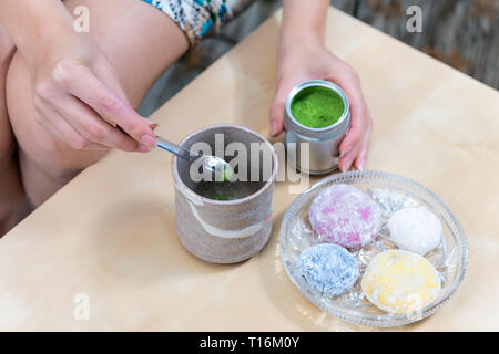 Green Matcha Teetasse und mochi Reiskuchen japanische Dessert wagashi Bunte daifuku mit Frau Mädchen Hand die Zubereitung von Tee Pulver Messlöffel auf Tisch Stockfoto