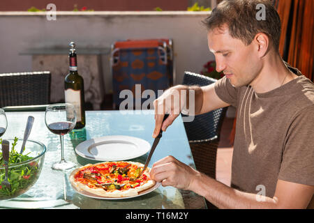 Junge glücklicher Mann die ganze kleine Pizza mit Messer auf der Terrasse Terrasse in Italien mit Flasche und Gläser Rotwein und Salat auf Glas Tisch Stockfoto