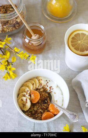 Frühstück mit Müsli mit Joghurt und Tee und forsythia Blumen auf einem Leinen Tischdecke. Im rustikalen Stil. Stockfoto