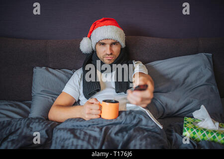Ruhe und ernsthaften jungen Mann sitzt auf dem Bett und gerade nach vorne zu schauen. Er ist mit der Decke zugedeckt. Kerl hält Tasse Tee und nutzt TV-Fernbedienung Stockfoto