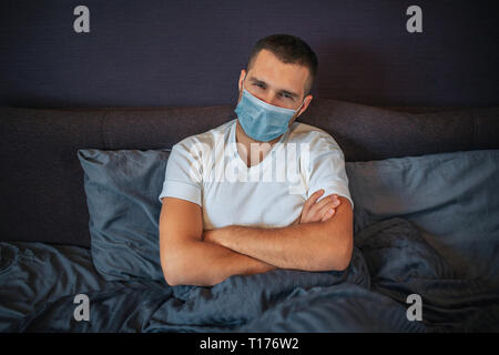 Ernsthafte junge Mann trägt Schutz Maske. Er sitzt auf dem Bett und schaut auf Kamera. Kerl hält die Hände gekreuzt. Er ist sehr ernst. Stockfoto