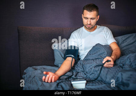 Ernsthafte junge Mann sitzt auf dem Bett und Maßnahmen seinen Blutdruck. Er schaut auf Gerät und verwendet es durch Zusammendrücken. Er ist entspannt. Kerl in im Schlafzimmer. Stockfoto