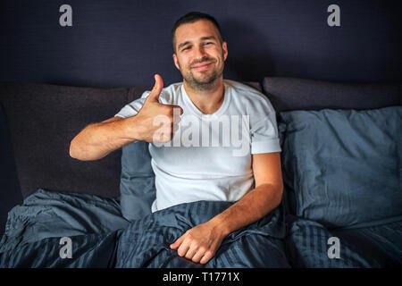 Positive junge Mann sitzt auf dem Bett und schaut auf Kamera. Er lächelt und zeigt beide Daumen nach oben. Kerl ist im Schlafzimmer. Stockfoto