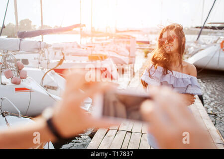 Schön und glücklich Modell steht und Posen vor der Kamera. Sie lächelt. Wind weht bis ihr Haar. Junge Frau trägt eine Sonnenbrille. Der Mensch nimmt ein Bild von Stockfoto