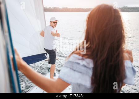 Junger Mann in Mütze und Brille steht auf Bogen der Yacht und erreicht mit der Hand zu brunette. Er schaut sie an. Sie steht und sieht ihn an. Modell hält an Stockfoto