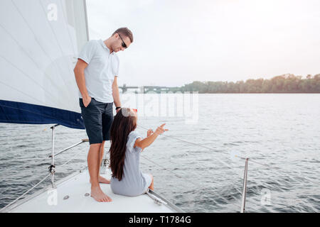 Paar ist Segeln auf der Yacht. Junge Frau sitzt auf Bogen und Punkte hinter. Sie schaut auf ihren Freund. Er lächelt und sieht sie an. Guy berührt brunette Stockfoto
