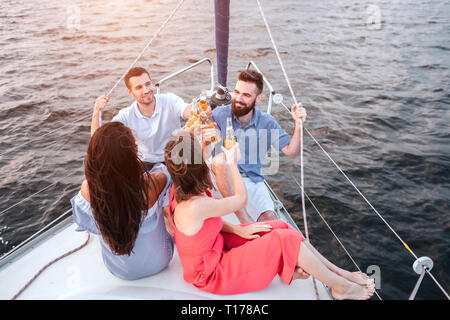 Junge Frauen sitzen mit Männer. Alle von Ihnen halten Flaschen mit Bier. Sie feiern. Kerl auf der linken Seite halten Seil mit Hand. Mann auf der rechten Seite ist Stockfoto