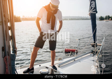 Ein weiteres Bild von oyung Mann steht auf der Yacht. Hr hält die Hände in den Taschen und nach unten schauen. Junge Mann hat Pullover auf Schultern und Kappe auf dem Kopf. Auch gibt Stockfoto