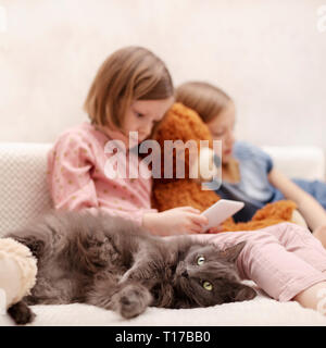 Zwei Mädchen (7 und 8 Jahre alt) gerade ein E-Buch und ein Smartphone zu Hause auf dem Sofa mit ihrer Katze. Selektive konzentrieren. Stockfoto