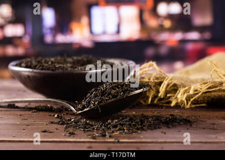 Viele Stücke von trockenen schwarzer Tee Earl Grey in einem grauen Keramik Schüssel auf Jute Tuch mit silbernen Löffel mit Restaurant im Hintergrund Stockfoto