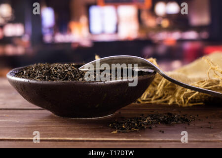 Viele Stücke von trockenen schwarzer Tee Earl Grey in einem grauen Keramik Schüssel auf Jute Tuch mit silbernen Löffel mit Restaurant im Hintergrund Stockfoto