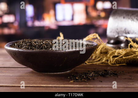 Viele Stücke von trockenen schwarzer Tee Earl Grey in einem grauen Keramik Schüssel auf Jute Tuch mit schwarzen Keramiktasse mit Restaurant im Hintergrund Stockfoto