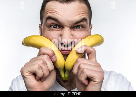 Ein ungewöhnlicher Mann im weißen Hemd hält zwei Bananen in der Nähe von seinem Gesicht und machen lustige Gesichter. Dieses vegane mag Obst und Gemüse zu essen. Er ist auch Stockfoto