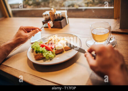 Nahaufnahme der Mann mit Messer und Gabel. Es gibt einen Teller leckere Waffel mit Gemüse und einer Tasse Tee. Tabelle steht vor windo Stockfoto