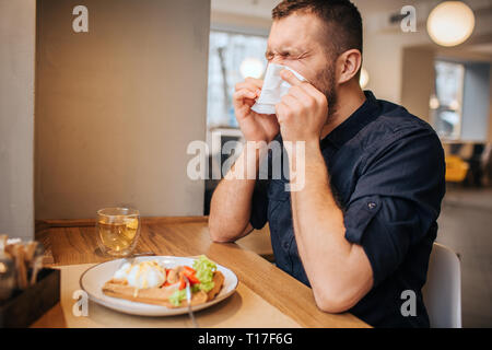 Kranken mann am Tisch sitzt und Niesen. Er ist für seine Nase mit weißen Serviette und halten die Augen geschlossen. Mann ist sehr krank Stockfoto
