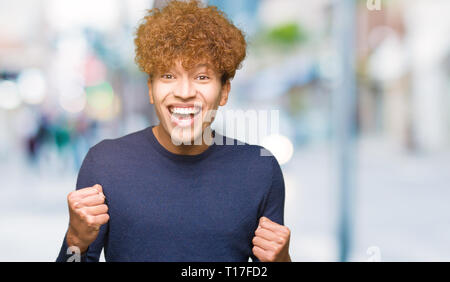 Junger stattlicher Mann mit afro Haar sehr glücklich und aufgeregt, Sieger Geste mit erhobenen Armen, lächelnd und Schreien für den Erfolg. Feier Konzept Stockfoto