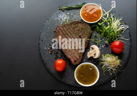 Lean gegrillte Steaks gewürzt mit Kräutern und Gewürzen serviert mit frischen grünen Sprossen, Tomaten und Dips oder Dressings von oben gesehen auf einem runden blac Stockfoto