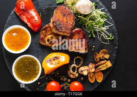 Serviert gegrilltes Schweinefilet mit sortierten gebratenes Gemüse, Tomaten, Soße und Salat Sprossen von Overhead auf einem schwarzen Brett gesehen Stockfoto