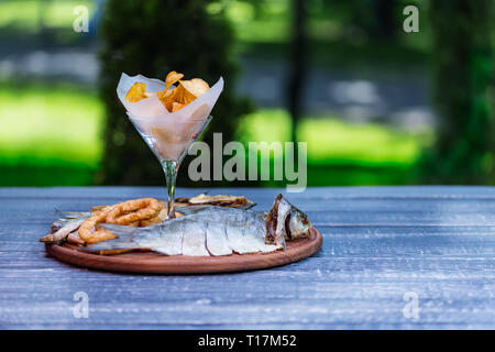 Snacks für Bier. Gesalzene, getrocknete Fische, Tintenfischringe und Kartoffelchips, auf dem Schneidbrett und grüner Sommer Hintergrund Stockfoto