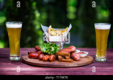 Wurst für Bier und zwei Gläser Bier. Würstchen, Pommes frites, Cracker, Ketchup und Sauce auf dem Schneidebrett, Holz- und Grüner Sommer Stockfoto