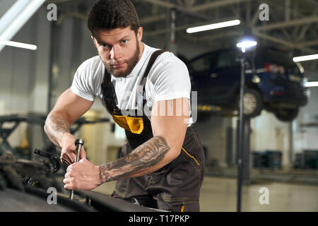 Muskulöse Mechaniker in Uniform Reparatur Auto in autoservise Station. Stattlich, brutale bärtigen Handwerker mit dunklen Haaren und Tattoos auf Hand Dirndl und Kamera. Stockfoto