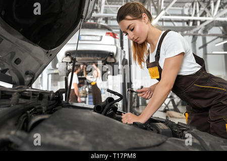 Schöne Mädchen mit Pferdeschwanz, tragen in Overalls und weißes T-Shirt arbeiten in Autoservice als Mechaniker. Hübsche, hübsche junge Frau zur Festsetzung unter dem Auto Motorhaube, mit Werkzeug, Ausrüstung. Stockfoto