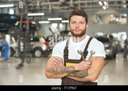 Schönen, muskulösen Mann im weißen T-Shirt, Overalls, arbeiten als Mechaniker in Autoservice. Brutal, tattoed Handwerker holding Druckminderer, posierend, an der Kamera schaut. Stockfoto