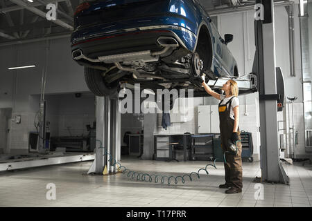 Professionelle Mechanikerin in der Nähe von Automobil auf Brücke angehoben, Beobachtung und Prüfung der Rad. Junge Frau zu Bremsscheiben. Mädchen tragen in Overalls. Stockfoto