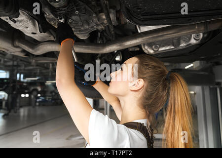 Junge Frau mit Werkzeug, zur Festsetzung Auto. Mädchen arbeiten Unterwagen von Automobile, Auto hob auf der Brücke. Mechanikerin in schwarz Handschuhe suchen, Reparatur Fahrzeug in Service Station. Stockfoto